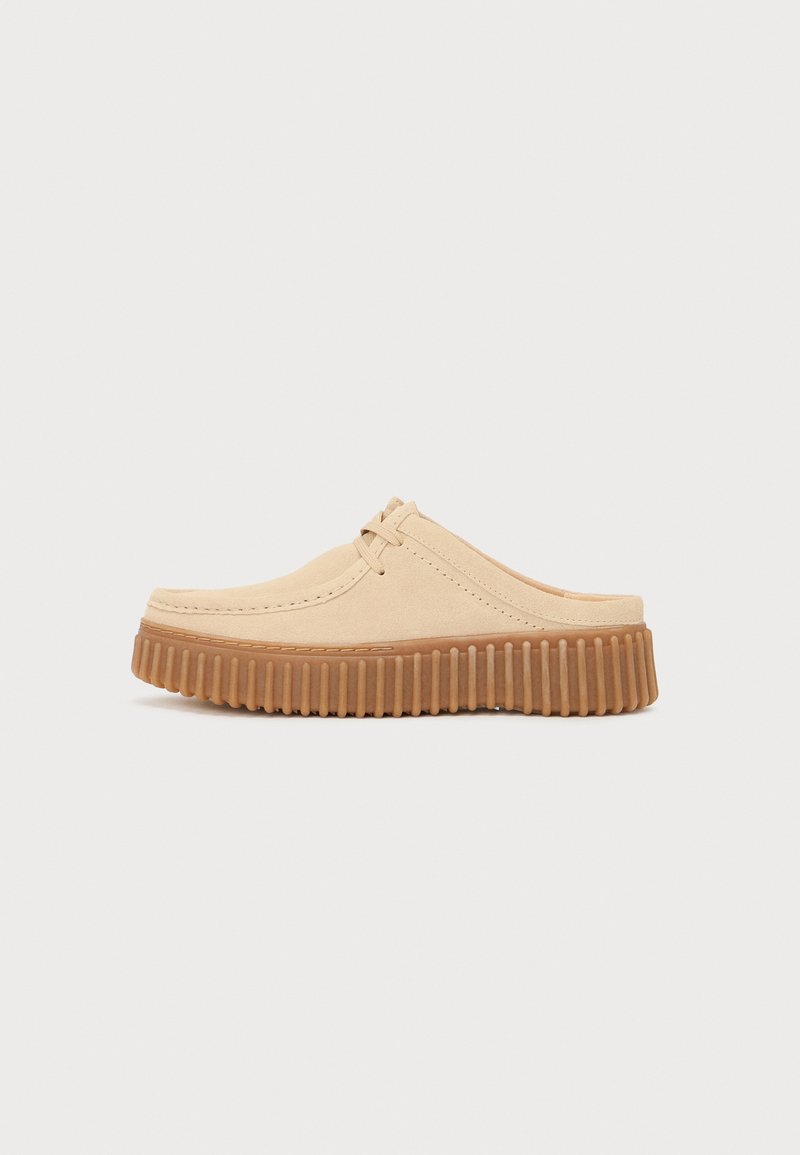 Beige suede clogs with a rounded toe, stitched detailing, and a textured rubber sole featuring vertical ridges for grip.