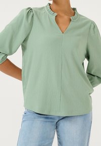 Blouse verte texturée avec un col à volants, manches trois-quarts. Présente un décolleté en V et un ourlet droit, portée avec un jean bleu clair.