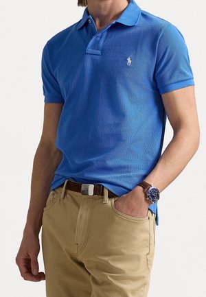 Homme portant un polo bleu avec un logo blanc, pantalon beige, ceinture marron et montre avec cadran bleu et bracelet en cuir marron, main dans la poche.