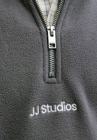 Pull en polaire gris avec un zip quart, doté d'une fermeture éclair argentée et d'un logo "JJ Studios" brodé en blanc sur la poitrine.