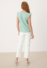 Světle zelený top z lněné směsi s volány na rukávech, v kombinaci s bílými zkrácenými džíny. Model stojí zády k fotoaparátu, aby předvedl detaily outfitu.