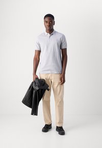 Lysegrå polo shirt med korte ærmer, beige bukser og sorte sneakers. Holder en sort læderjakke i venstre hånd.