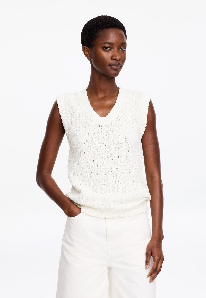 Femme aux cheveux courts portant un pull blanc texturé sans manches et un pantalon blanc assorti, debout avec une main dans la poche.