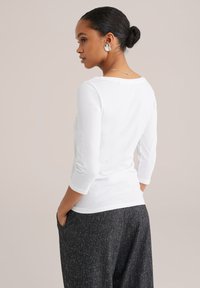 Frau mit einem Dutt trägt ein weißes Langarmshirt, dunkle strukturierte Hose, silberne Ohrringe und eine dünne silberne Halskette und steht mit den Händen in den Taschen.