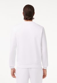 Homme aux cheveux foncés mouillés portant un sweat-shirt blanc uni et un pantalon blanc, vu de dos sur un fond blanc.