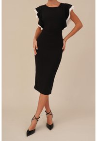 Svart figurnära midiklänning med flygande ärmar och vit kantning. Slät textur, minimalistisk design. Åtsittande tillsammans med svarta högklackade sandaler.