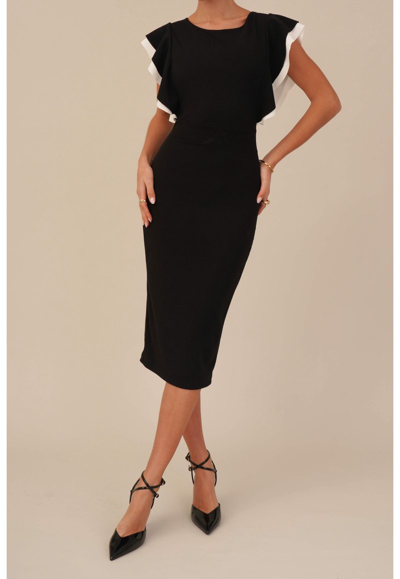 Svart figurnära midiklänning med flygande ärmar och vit kantning. Slät textur, minimalistisk design. Åtsittande tillsammans med svarta högklackade sandaler.