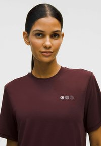 Sötét hajú nő, aki lófarokba fogta a haját, bordó színű pólót visel, amelyen kicsi fehér logók találhatók a mellkasán, egy sima világos háttér előtt.