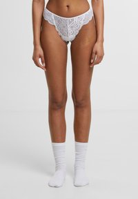 Witte kanten string met golvende randen, versierd met bloemenpatronen. Model draagt witte geribbelde enkelsokken. Achtergrond is eenvoudig.