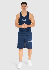 Modelo masculino vestido con una camiseta deportiva sin mangas de color azul marino y pantalones cortos, con zapatillas blancas, de pie con las manos entrelazadas contra un fondo gris claro.