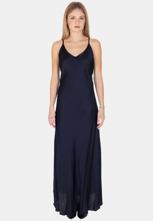 Giovane donna che indossa un lungo abito di raso blu navy con spalline sottili, in piedi davanti a uno sfondo chiaro e uniforme.