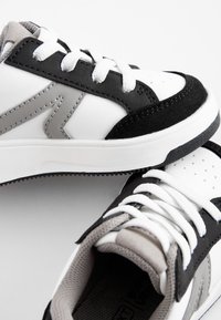 Zapatillas deportivas en negro, blanco y gris con cuero sintético, detalles de malla, suelas texturizadas y cordones blancos. Presentan una caja de dedos perforada.
