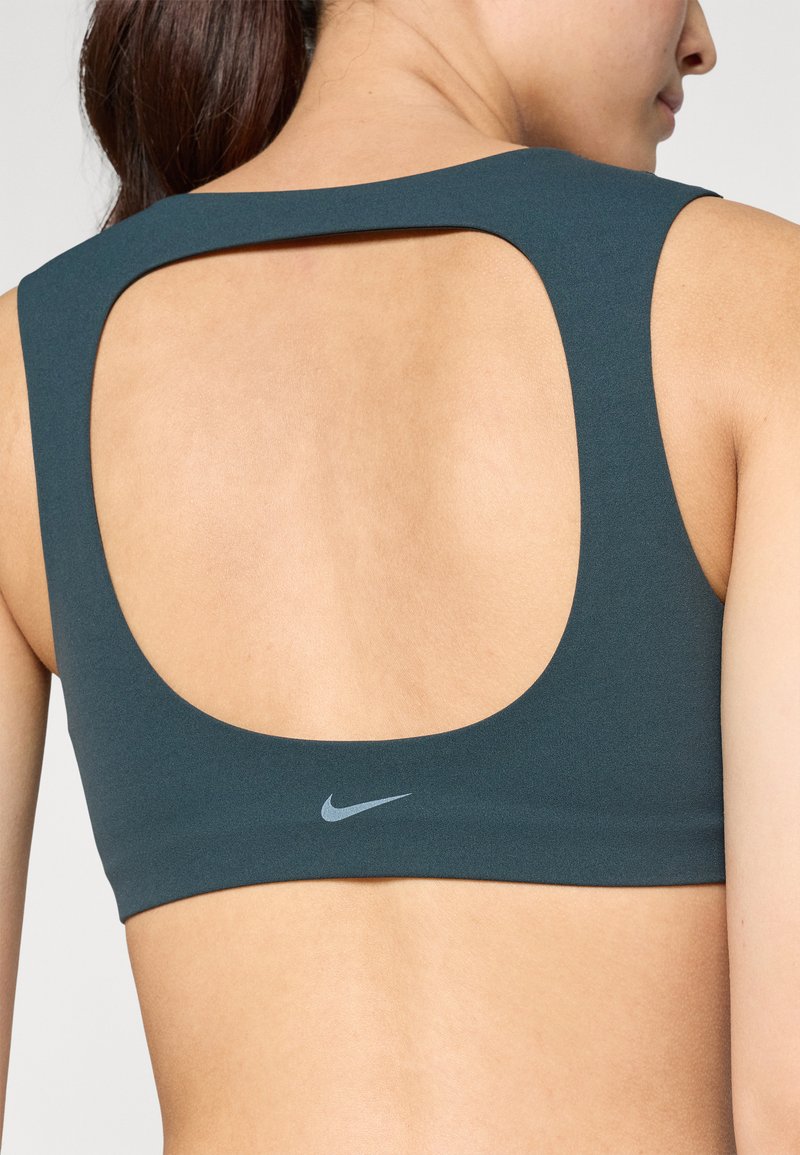 Donker teal sportbeha met een open rugontwerp. Heeft een soepele stof en een klein grijs Nike-logo gecentreerd op de band.