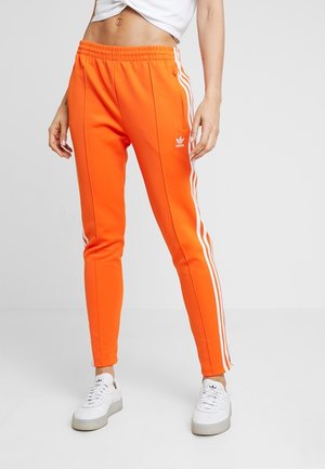 Persona con pantalones de chándal Adidas naranja brillante con rayas blancas en los lados y zapatillas blancas sobre un fondo liso.