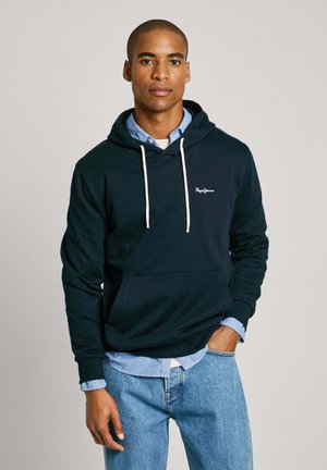 Dunkelblaues Kapuzensweatshirt mit weißen Kordeln, Fronttasche und gesticktem Logo. Trägt man über einem hellblauen Hemd mit Kragen.