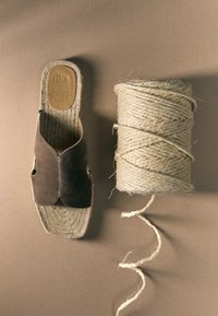 Une sandale en daim marron unique avec une semelle tissée placée à côté d'une grande bobine de corde blanche épaisse sur un fond beige.