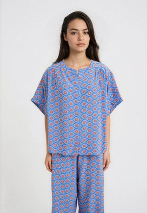 Frau mit langen dunklen Haaren, die ein passendes lockeres blaues Oberteil und Hose mit rotem und weißem Blumenmuster trägt, steht vor einfarbigem Hintergrund.