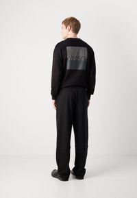 Zwarte sweatshirt met een groot matte logo op de achterkant, gecombineerd met losvallende zwarte broek. Beide items zijn gemaakt van zachte stof.
