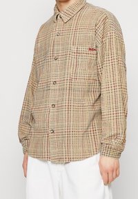 Beige geruite overhemd met lange mouwen, voorzien van rode en marineblauwe lijnen, knoopsluiting aan de voorkant en een linkerborstzak met een rood logo. Gekreukte textuur.