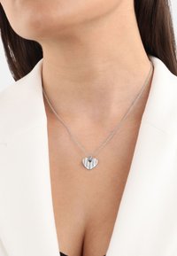 Collar de corazón de plata con un diseño texturizado y cadena. Presenta rayas y acentos brillantes, expuesto sobre un fondo claro.