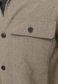 Giacca camicia in misto lana marrone con tasca frontale e bottoni neri. Il tessuto ha una texture morbida e un delicato motivo a trama.