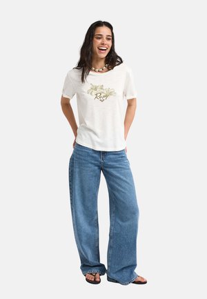 Vrouw glimlacht en staat, draagt een wit gebloemd Roxy T-shirt, wijde blauwe jeans, schelpenketting en zwarte sandalen tegen een egale achtergrond.