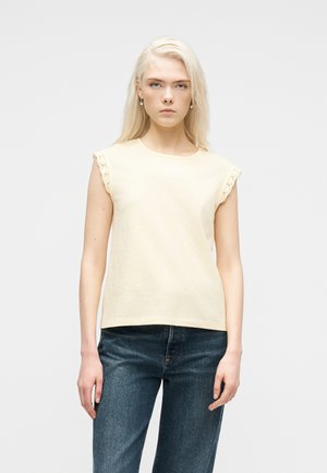 Jeune femme aux longs cheveux blonds portant un haut crème sans manches avec bordure en dentelle et un jean bleu, debout devant un fond clair uni.