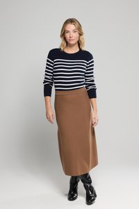 Vrouw met blond haar, gekleed in een marineblauw en witte gestreepte trui, een bruine midi-rok en zwarte hoge hakken, staande tegen een effen lichtgrijze achtergrond.