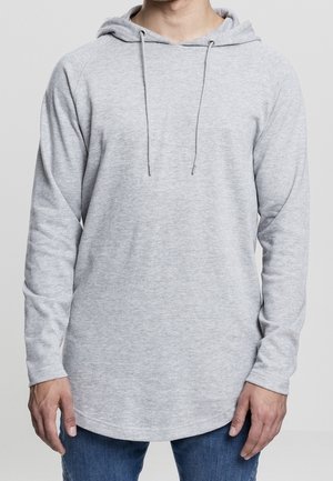 Mand iført en lysegrå hættetrøje med snore og lange ærmer, kombineret med blå jeans, stående mod en ensfarvet baggrund.