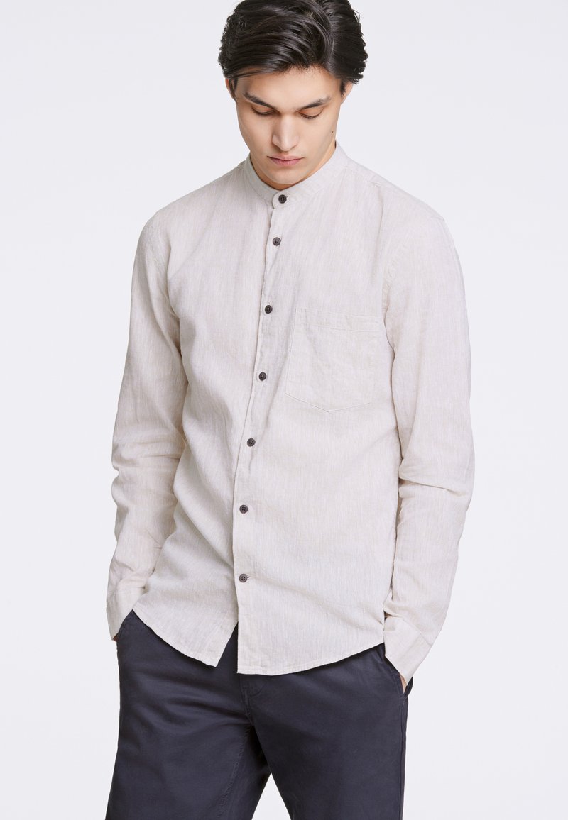 Camisa de hombre de manga larga tipo botonera en tejido beige claro, con un diseño sin cuello, bolsillo frontal y botones marrones oscuros.