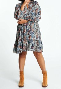 Personne portant une robe longue à manches longues à motif floral dans des tons bleu, marron et blanc, associée à des bottines en daim couleur camel, debout les bras croisés.