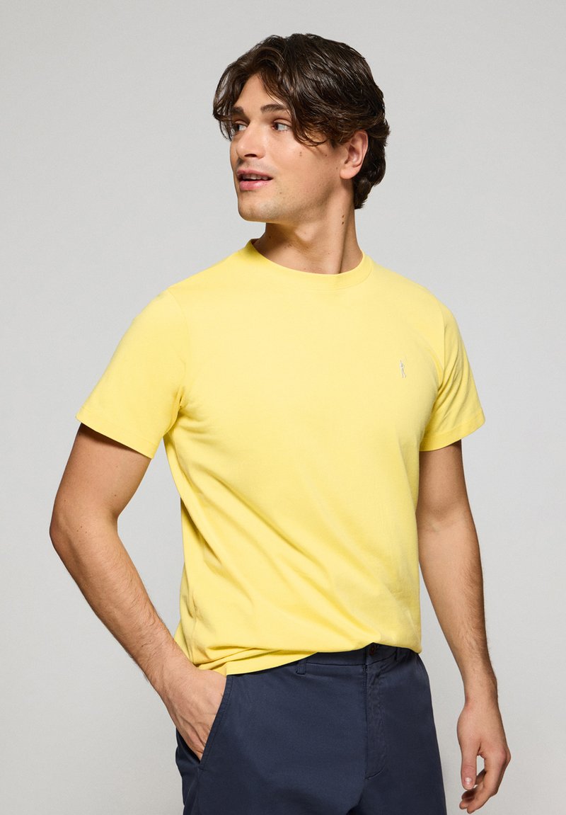 Camiseta de algodón amarilla con mangas cortas, cuello redondo y un pequeño logo bordado en el pecho. El modelo lleva puesto un pantalón marino.
