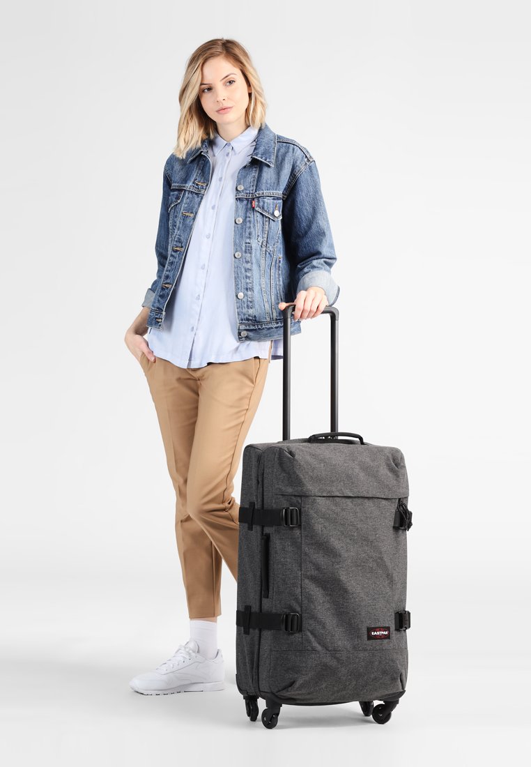 Gray fabric rolling suitcase with four wheels, retractable handle, side straps, and red logo patch. Casual outfit shown for context.