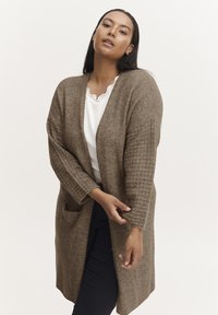 Brown knitted cardigan with open front, textured sleeves, and side pockets. Worn over a white top and dark pants. Smooth, warm fabric.