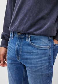 Jeans din denim de culoare albastru deschis, cu un design ajustat, având cinci buzunare, închidere cu buton și o textură ușor decolorată. Asortate cu o cămașă lungă și închisă la culoare.