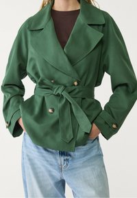 Grøn, dobbeltradet trench coat med bælte i taljen, klassiske slag og store knapper, sat sammen med lyseblå jeans. Glat stof.