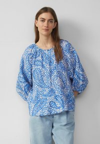 Blaue gemusterte Bluse mit Ballonärmeln, rundem Halsausschnitt und leicht strukturiertem Stoff. Kombiniert mit hellblauen Jeans.