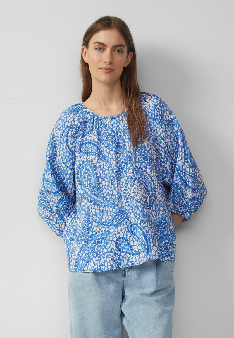 Blauwe blouse met patroon, ballonmouwen, ronde halslijn en lichte textuur. Gecombineerd met lichtblauwe jeans.