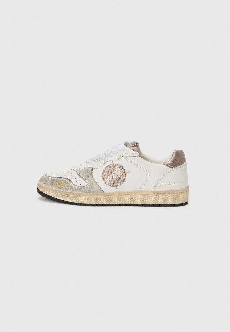 Sneaker bianco con accenti in suede grigio, patch rotondo con logo, testo dorato e lacci bianchi. Presenta una suola in gomma testurizzata e una parte superiore in pelle liscia.