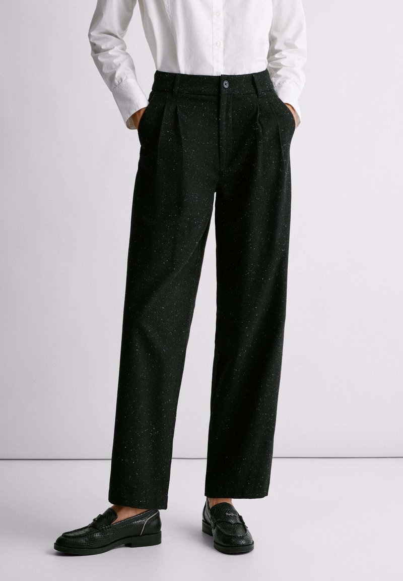 Pantaloni neri, strutturati e plissettati, con una vestibilità ampia e chiusura con bottone, caratterizzati da piccole macchie di colore sparse. Indossati con scarpe nere.