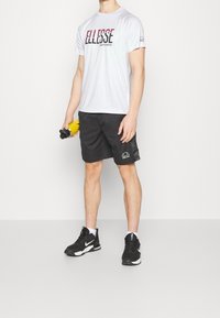 Camiseta deportiva blanca con el logo "Ellesse" en rojo y negro, combinada con pantalones cortos negros que llevan un logo. Zapatillas negras con detalles en blanco.