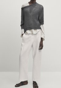 Grijze gebreide trui met een asymmetrische halslijn over een wit gestreefd shirt, gecombineerd met witte linnen wijde broek en zwarte sandalen.