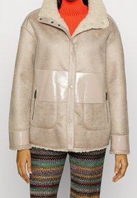 Beige Jacke mit einem Kragen aus Kunstschafsfell, glänzender Brustpartie und mit Reißverschlüssen versehenen Seitentaschen. Strukturierter Stoff mit glatten Akzenten. Kombiniert mit gemusterten Hosen.