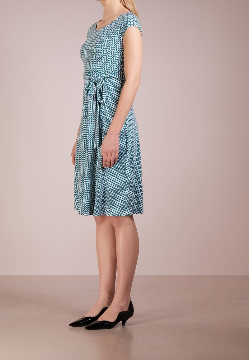 Femme portant une robe bleue à motifs mi-longue avec un lien à la taille et des chaussures noires à talons bas, debout de profil sur un fond uni.