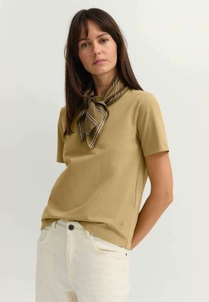 Frau mit glattem braunem Haar, die ein kurzärmeliges, beige Oberteil, einen gestreiften Schal um den Hals gebunden und weiße Hosen vor einem einfarbigen Hintergrund trägt.