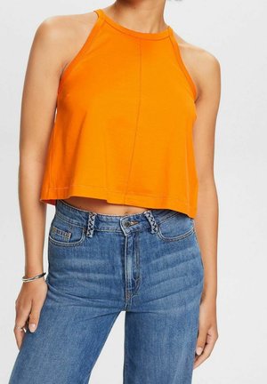 Woman wearing a sleeveless orange crop top and high-waisted blue jeans, with a silver bracelet and rings on her left hand.