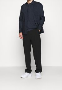 Marineblå skjorte med knapper og brystlomme over en mørk t-shirt, parret med sorte bukser og hvide sneakers. Minimalistisk design og pasform.