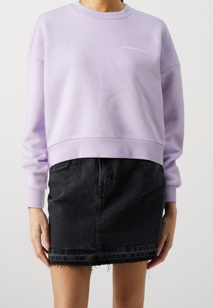 Femme portant un sweat-shirt col rond violet clair avec un petit texte et une mini-jupe en jean noire avec un ourlet effiloché, debout devant un fond uni.