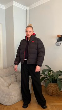 Črna puhovka z rdečo notranjostjo, z zadrgo in logotipom blagovne znamke na prsih. Kombinirana z ohlapnimi črnimi hlačami in masivnimi črnimi čevlji.