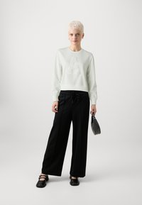 Hanorac verde deschis cu un design texturat al literei "A", asortat cu pantaloni negri evazați și pantofi negri. Ține în mână o poșetă mică neagră.